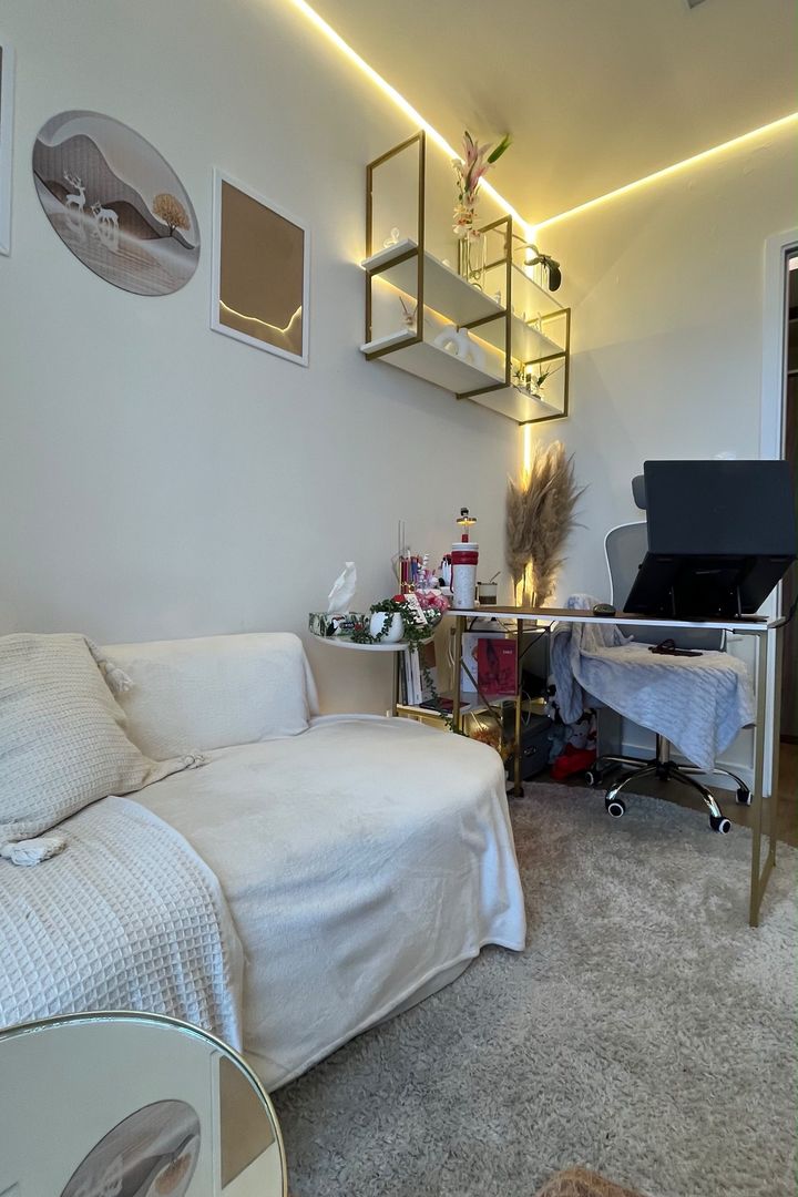 Sofá com manta, quadros na parede, prateleiras douradas e mesa de trabalho ao fundo.